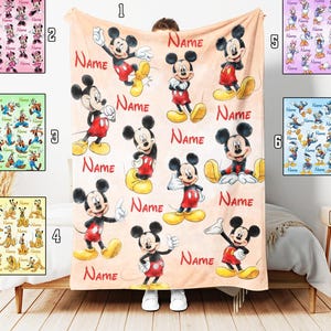 May include: A beige blanket with multiple Mickey Mouse images and the word "NAME" in red. Other blankets with Disney characters are visible. The blanket is displayed over a person. The blanket is approximately 150 cm x 200 cm.