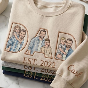 May include: A stack of beige, gray, green, and navy blue sweatshirts. The top sweatshirt features the word "DAD" with embroidered family portraits and "EST. 2022". The sleeve is embroidered with the name "Carl".