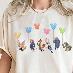 May include: Cream-colored t-shirt featuring a whimsical illustration of cartoon characters holding Mickey Mouse-shaped balloons in various colors. The characters include a fox, lion, rabbit, and others. The design is centered on the shirt.