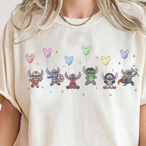 May include: Cream-colored t-shirt featuring a colorful graphic of cartoon characters dressed as superheroes, each holding a Mickey Mouse-shaped balloon. The characters include Iron Man, Captain America, Deadpool, Hulk, Wonder Woman, and Thor.
