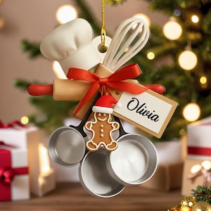Peut inclure: Ornement de Noël représentant des ustensiles de cuisine miniatures : toque de chef, fouet, rouleau à pâtisserie et tasses à mesurer, noués avec un ruban rouge. Un bonhomme en pain d'épices et une étiquette avec le nom "Olivia" sont également inclus.