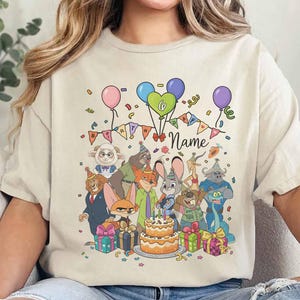 Puede incluir: Camiseta beige con un dise&ntilde;o de cumplea&ntilde;os colorido. Presenta personajes de dibujos animados, globos, confeti, una tarta y la palabra "Name". El dise&ntilde;o incluye una pancarta que dice "Birthday".