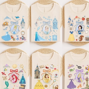May include: Six ivory-colored t-shirts with different princess-themed designs. Each shirt features a princess portrait surrounded by related icons, such as castles, dresses, and accessories. The shirts are neatly folded on woven mats.