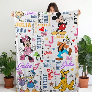 May include: White blanket featuring various Disney characters, including Mickey Mouse, Minnie Mouse, Goofy, Donald Duck, and Pluto. The name "Julia" is repeated throughout the design in different colors and fonts. The blanket is held up by a person.