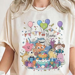 May include: Cream-colored t-shirt featuring a colorful birthday scene with cartoon sea creatures, balloons, and a cake. The design includes the word "Name" and a banner that says "Happy Birthday". The shirt is a casual, comfortable style.
