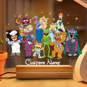 May include: A decorative night light featuring a clear acrylic panel with a colorful illustration of Muppet characters. The base is made of wood and has the text "Custom Name" on the front. The light is on, illuminating the characters.