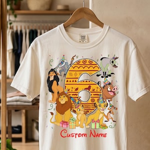 May include: A cream-colored short-sleeve t-shirt featuring a large orange number six decorated with tribal patterns. The graphic includes various animated lion characters wearing party hats, colorful gift boxes, and the red text Custom Name.