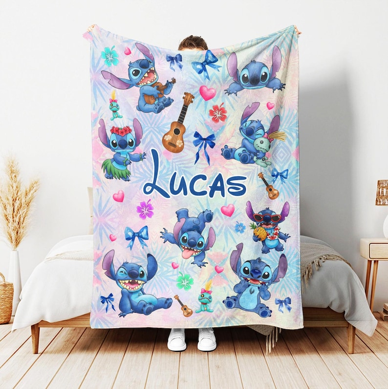 Op de afbeelding: Een gepersonaliseerde deken met het personage Stitch uit Lilo & Stitch. Het ontwerp bevat meerdere afbeeldingen van Stitch, ukeleles en bloemaccenten op een pastelkleurige achtergrond. De naam "LUCAS" staat prominent in blauwe letters.