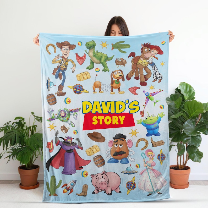 Puede incluir: Una manta de forro polar azul claro con un patr&oacute;n colorido de varios personajes animados, incluyendo un vaquero, un guardi&aacute;n espacial, un dinosaurio, un cerdo y una figura de patata, con el texto "DAVID'S STORY" en letras amarillas y rojas.