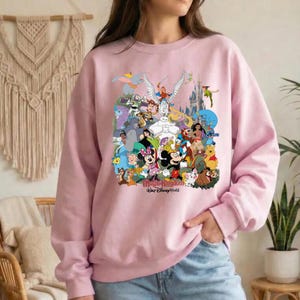 Puede incluir: Sudadera rosa claro con un collage colorido de personajes de Disney, incluyendo Mickey Mouse, Minnie Mouse y Winnie the Pooh. Las palabras "Magic Kingdom Walt Disney World" est&aacute;n impresas en la parte inferior del dise&ntilde;o.