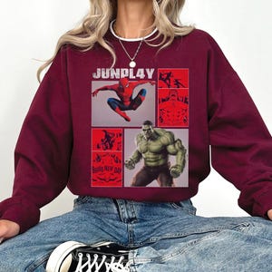 Op de afbeelding: Bordeaux sweatshirt met een grafisch ontwerp met de tekst "JUNPLAY" en afbeeldingen van Spider-Man en Hulk. Het ontwerp bevat rode panelen met illustraties en de tekst "BRAND NEW DAY".