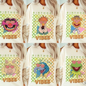 May include: Ivory t-shirts with Muppet characters wearing sunglasses. Each shirt has the words "BIRTHDAY VIBES" in a retro font, set against a checkered and geometric background. The shirts are long-sleeved.