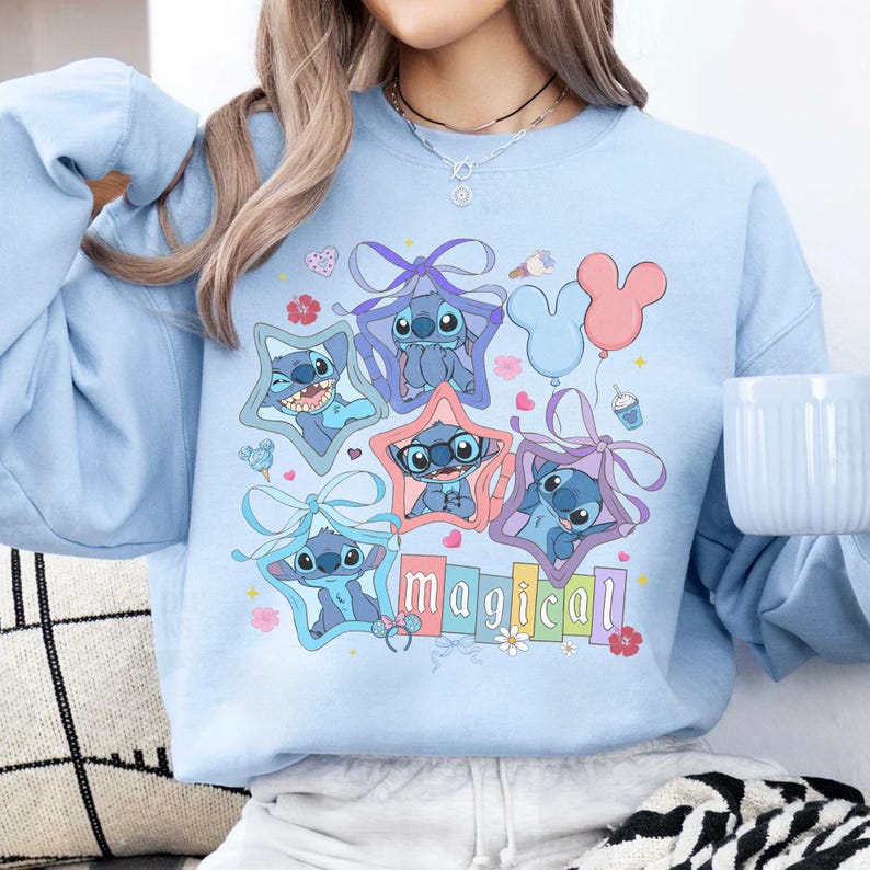 Może przedstawiać: Jasnoniebieska bluza z kolorową grafiką Stitcha z Lilo & Stitch. Projekt zawiera postacie Stitcha w ramkach w kształcie gwiazd, balony i słowo "magical" w tęczowych kolorach. Zabawny i fantazyjny design.
