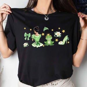 Peut inclure: T-shirt noir avec un motif vert sur le th&egrave;me de la princesse. Le motif comprend une princesse, une grenouille avec une couronne, un escarpin, un n&oelig;ud et des &eacute;toiles. La princesse porte une robe verte et a les cheveux attach&eacute;s en chignon.
