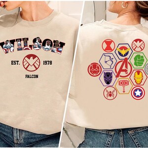 Puede incluir: Sudadera beige con la palabra "WILSON" en estilo c&oacute;mic, con ilustraciones de personajes. Debajo, un logotipo de escudo rojo con "EST. 1978 FALCON". La parte trasera muestra varios logotipos de superh&eacute;roes en formas hexagonales coloridas.