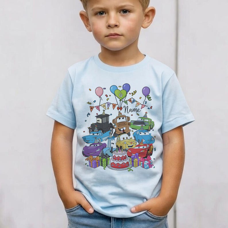 Peut inclure: Un t-shirt bleu clair avec un motif de dessin anim&eacute; de personnages de Cars, des ballons, un g&acirc;teau d'anniversaire et des bo&icirc;tes cadeaux. Le mot "Name" est &eacute;crit en cursive. Le t-shirt est pour enfants.