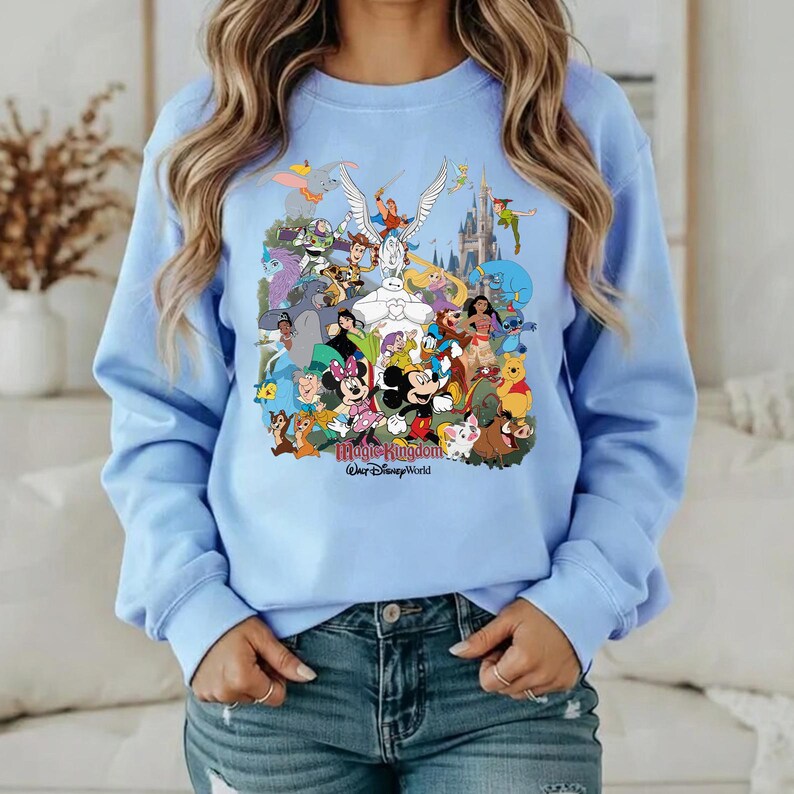 Puede incluir: Sudadera azul claro con un gr&aacute;fico colorido de varios personajes de Disney, incluyendo Mickey Mouse, Minnie Mouse y Winnie the Pooh. El texto "Magic Kingdom" y "Walt Disney World" est&aacute; impreso debajo.