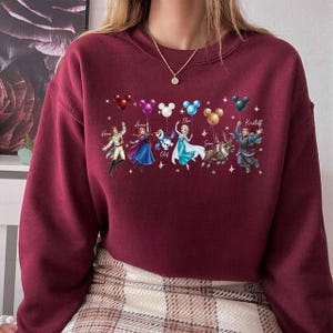 May include: Burgundy crewneck sweatshirt featuring a graphic of characters from a popular animated film. The characters are holding Mickey Mouse-shaped balloons in various colors. Names of the characters are printed above them.