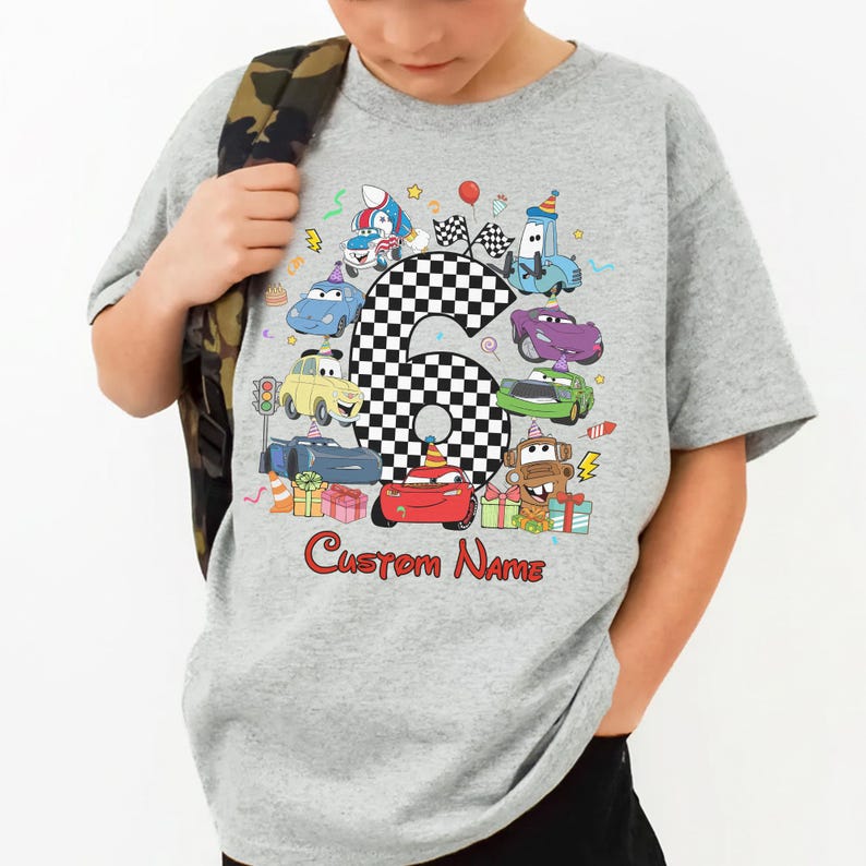 May include: A grey t-shirt with a vibrant cartoon car design, including the number 6 in a chequered pattern. The design features various cars, gifts, and the text "Custom Name" in red. The t-shirt is worn by a child.