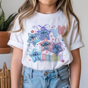 Może przedstawiać: Biała koszulka z kolorową grafiką postaci Stitch z filmu Lilo & Stitch. Stitch jest przedstawiony w ramkach w kształcie gwiazd z kokardkami, otoczony sercami, kwiatami i balonami. Słowo "magical" jest napisane kolorowymi literami.