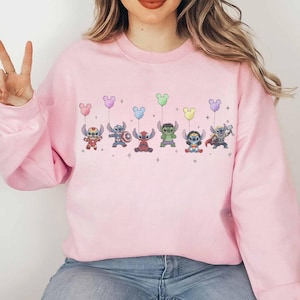 May include: A light pink sweatshirt featuring a line of cartoon characters holding Mickey Mouse-shaped balloons in various colors. The characters are dressed as superheroes. The sweatshirt is made of a soft material.