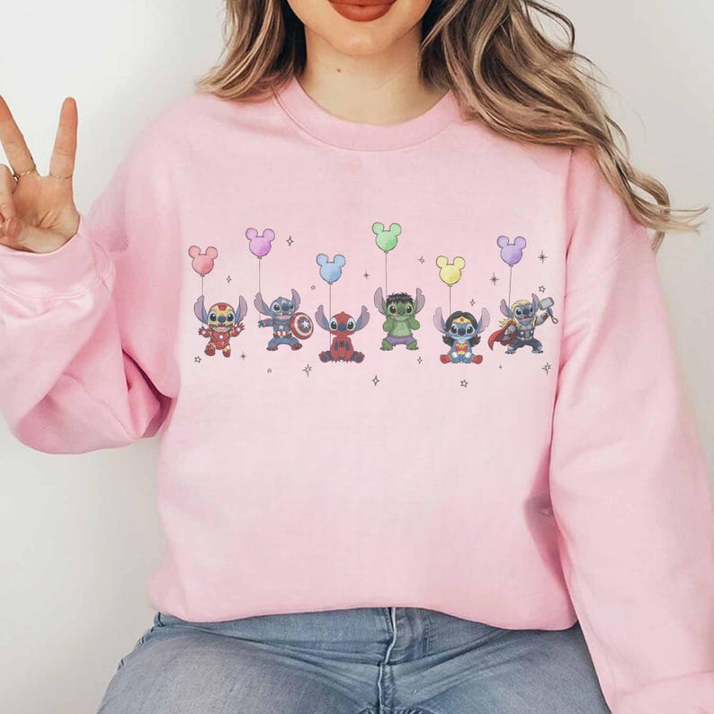May include: A light pink sweatshirt featuring a line of cartoon characters holding Mickey Mouse-shaped balloons in various colors. The characters are dressed as superheroes. The sweatshirt is made of a soft material.
