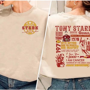 May include: Beige sweatshirt featuring a graphic of Tony Stark with text. The front of the sweatshirt has a gold and red Stark Industries logo. The back of the sweatshirt has a red graphic with text about Tony Stark, including "I am Iron Man."