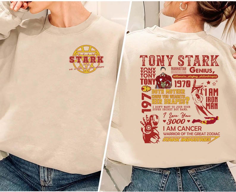 May include: Beige sweatshirt featuring a graphic of Tony Stark with text. The front of the sweatshirt has a gold and red Stark Industries logo. The back of the sweatshirt has a red graphic with text about Tony Stark, including "I am Iron Man."