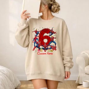 May include: A beige crewneck sweatshirt featuring a large red number six with a spiderweb pattern, surrounded by multiple illustrations of Spider-Man wearing party hats, colourful gift boxes, and the red text Custom Name printed below.