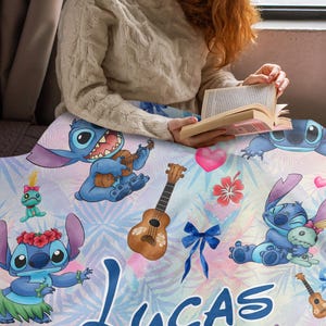 Op de afbeelding: Een gepersonaliseerde deken met een blauw stripfiguur dat ukulele speelt, omringd door andere figuren, ukeleles en tropische bloemen. De naam "LUCAS" staat in blauwe letters onderaan. De deken heeft een lichtblauwe en roze achtergrond.