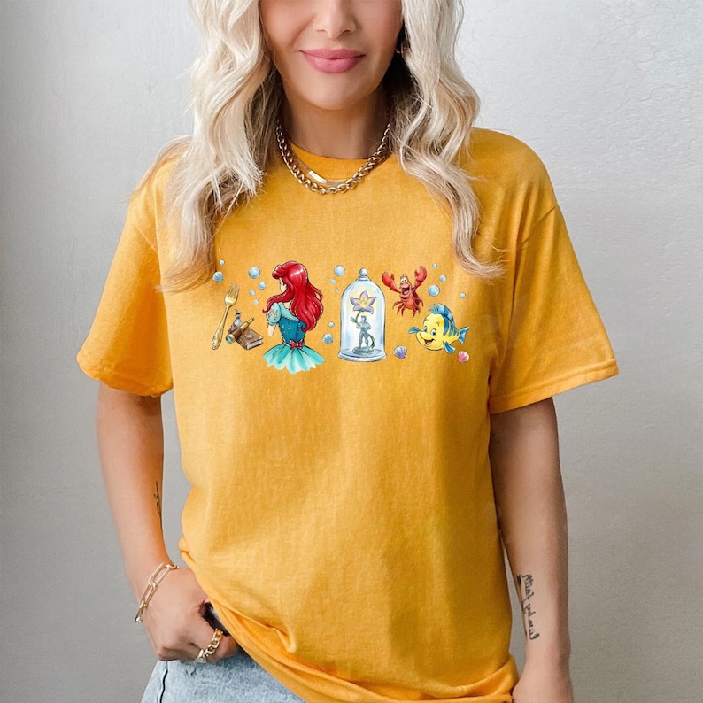 Peut inclure: T-shirt jaune moutarde avec un graphisme color&eacute; de personnages d'un film d'animation populaire. Le motif comprend une sir&egrave;ne rousse, un crabe, un poisson et d'autres &eacute;l&eacute;ments connexes. Le t-shirt a des manches courtes et un col rond.