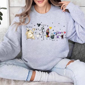 K&ouml;nnte beinhalten: Ein hellgraues Sweatshirt mit einer farbenfrohen Grafik von Figuren aus einem beliebten Zeichentrickfilm. Die Grafik zeigt Charaktere wie Jack Skellington, Sally und Oogie Boogie. Die Namen der Charaktere sind dar&uuml;ber gedruckt.