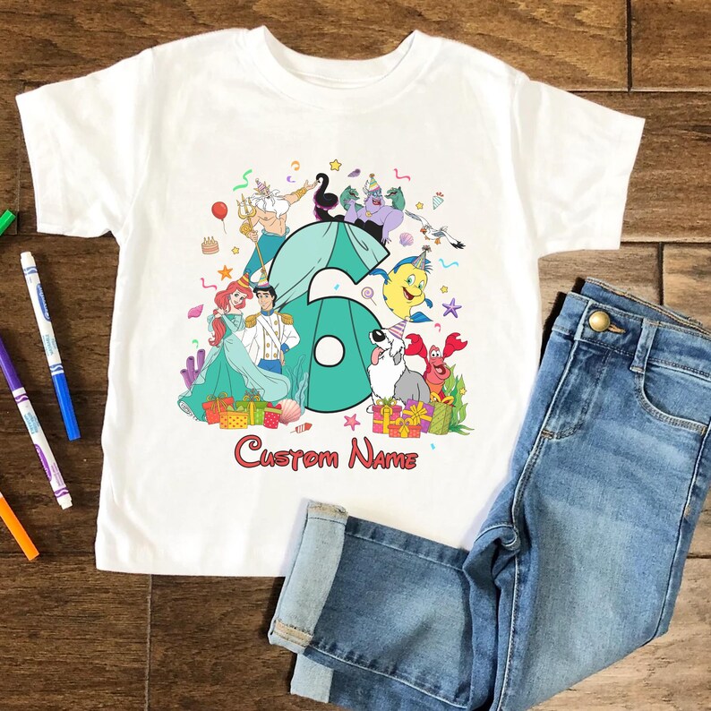 K&ouml;nnte beinhalten: Ein wei&szlig;es Kurzarm-T-Shirt mit einer gro&szlig;en t&uuml;rkisfarbenen Zahl sechs, umgeben von bunten Zeichentrickfiguren aus Arielle, die Meerjungfrau, darunter Arielle, Prinz Erik, Fabius, Sebastian, Triton und Ursula, mit dem Text Custom Name darunter.