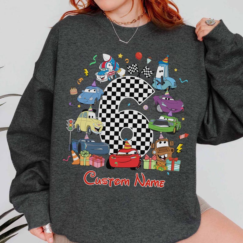 May include: Dark grey sweatshirt with a vibrant cartoon car design, including the number 6 in a chequered pattern. The design features car characters, party decorations, and the text "Custom Name" in red.
