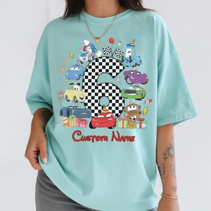 May include: A pale blue t-shirt with a vibrant graphic of cartoon cars, a chequered number six, and the words "Custom Name" in red. The design includes birthday-themed elements like a cake and presents.