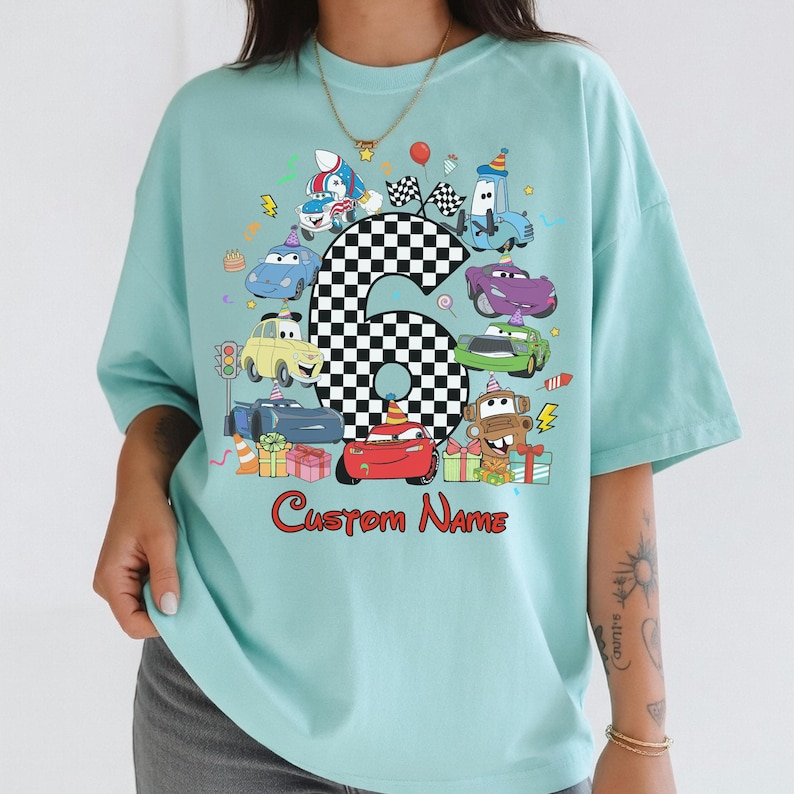 May include: A pale blue t-shirt with a vibrant graphic of cartoon cars, a chequered number six, and the words "Custom Name" in red. The design includes birthday-themed elements like a cake and presents.