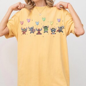 May include: Mustard yellow t-shirt featuring cartoon characters dressed as superheroes, each holding a Mickey Mouse-shaped balloon. The characters include Stitch, Captain America, and Thor. The design is printed in vibrant colors.