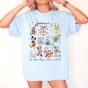 Puede incluir: Camiseta azul claro con un dise&ntilde;o colorido de Disney. El dise&ntilde;o incluye personajes como Mickey y Minnie Mouse, y lugares emblem&aacute;ticos. El texto dice "Walt Disney World", "2026" y "The Most Magical Place on Earth".