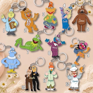 Könnte beinhalten: Eine Sammlung von zwölf Muppet-Schlüsselanhängern aus Acryl, wobei jede nummerierte Figur einen personalisierten Namen auf ihrer Kleidung oder ihrem Zubehör trägt und an einem silberfarbenen Metallring mit Kette befestigt ist.