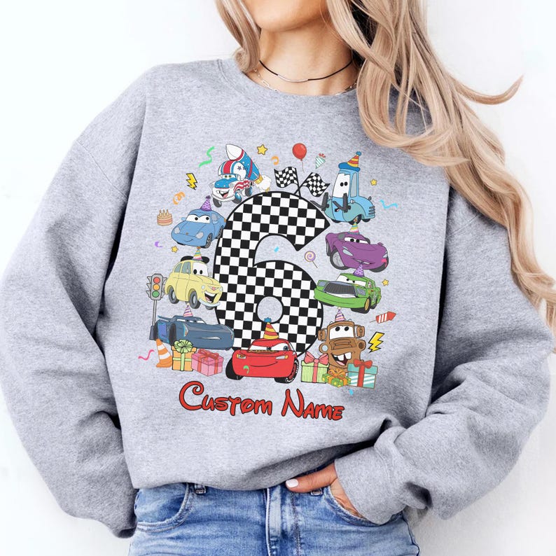 May include: A grey sweatshirt with a colourful car-themed design for a sixth birthday. The design includes a large number 6 in a chequered pattern, cartoon cars, and the text "Custom Name" in red script.