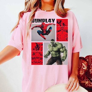 Op de afbeelding: Lichtroze t-shirt met een stripboekontwerp. Het ontwerp bevat Spider-Man, Hulk en andere illustraties in rood en wit. Het woord "JUNPLAY" staat bovenaan het t-shirt.