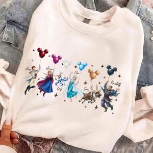 May include: White sweatshirt featuring a colorful graphic of characters from the movie Frozen, each holding a Mickey Mouse-shaped balloon. Characters include Anna, Elsa, Olaf, Kristoff, and others. The names of the characters are printed above them.
