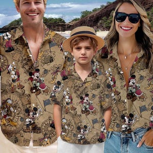 May include: Three matching tan button-down shirts with a cartoon pirate-themed design featuring Mickey Mouse, Donald Duck, and other characters. The shirts are worn by three people on a beach.