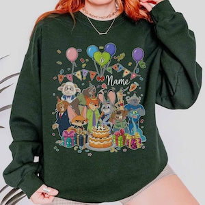 Puede incluir: Sudadera verde oscuro con un dise&ntilde;o de fiesta de cumplea&ntilde;os. Presenta personajes de dibujos animados, globos y una tarta de dos pisos. La palabra "Name" est&aacute; escrita en cursiva. Los regalos y confeti completan el dise&ntilde;o festivo.