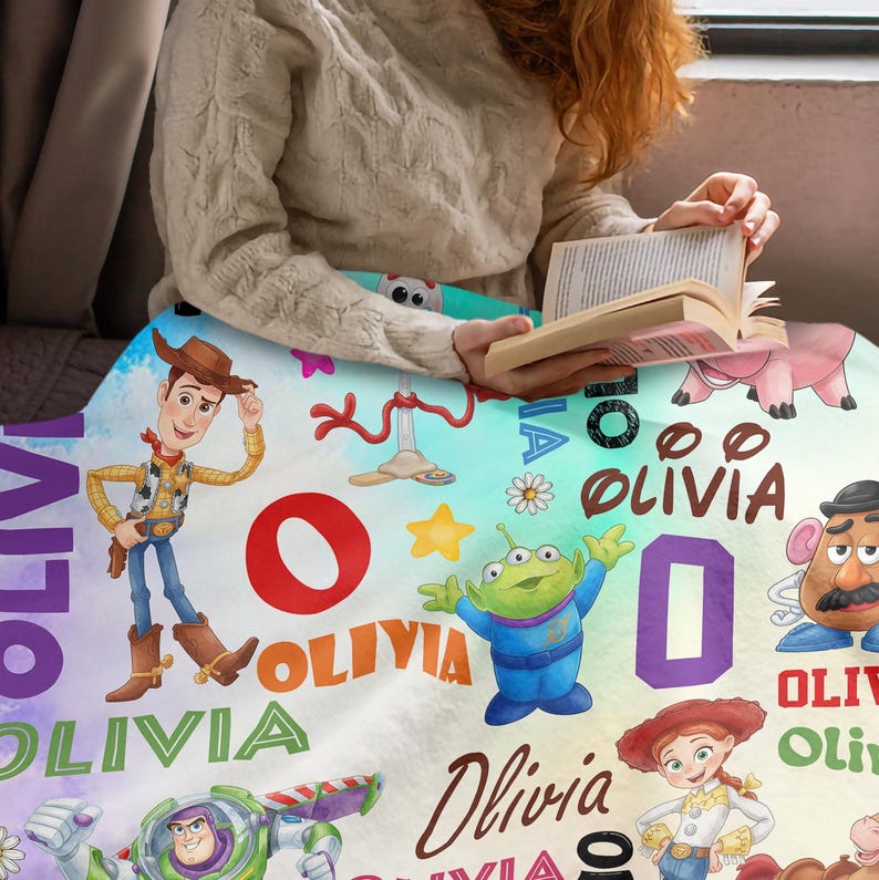 Op de afbeelding: Een zachte fleece deken met een kleurrijke Toy Story-karakterprint met Woody, Buzz Lightyear, Jessie en Mr. Potato Head. Het ontwerp bevat de naam Olivia die in verschillende lettertypes en kleuren wordt herhaald, naast sterren.