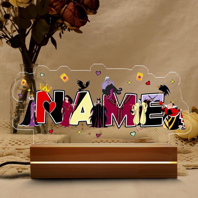 May include: A personalized LED night light featuring the word "NAME" with a Disney Villains theme. The clear acrylic design is illuminated by a wooden base with a warm glow. The letters are in black, red, and yellow, with character illustrations.