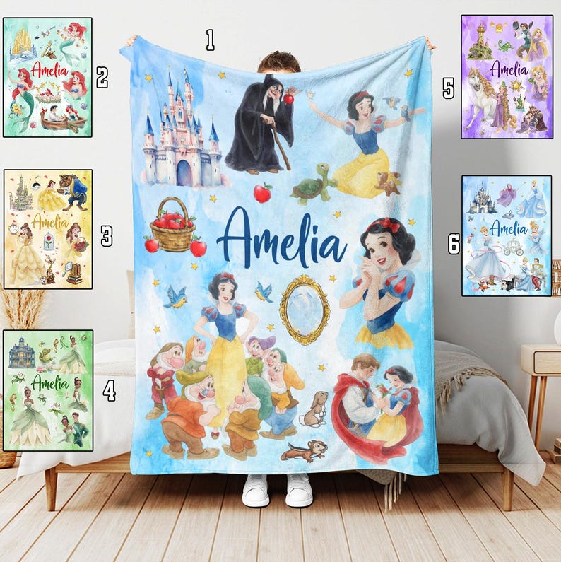 May include: A blue blanket with a Snow White theme, featuring illustrations of the princess, a castle, and apples. The name "Amelia" is written in blue script. The blanket is displayed on a bed, with a watercolor design.