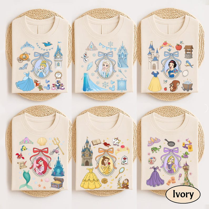 May include: Six ivory-colored t-shirts with different princess-themed designs. Each shirt features a princess portrait surrounded by related icons, such as castles, dresses, and accessories. The shirts are neatly folded on woven mats.