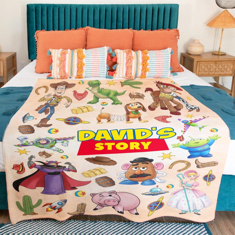 Puede incluir: Una manta de forro polar beige con un estampado colorido de personajes de Toy Story, incluyendo a Woody, Buzz Lightyear, Rex y el Sr. Patata, con el texto "DAVID'S STORY" impreso en grandes letras amarillas y rojas en el centro de la tela.