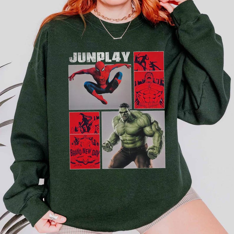 Op de afbeelding: Donkergroene sweatshirt met een grafisch ontwerp met Spider-Man en de Hulk. Het woord "JUNPLAY" staat bovenaan gedrukt, met de zin "BRAND NEW DAY" onderaan. Het ontwerp bevat rode en grijze stripboekpanelen.